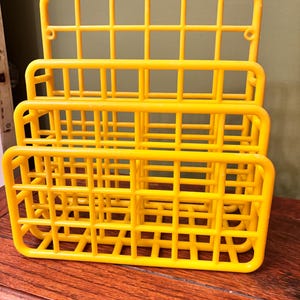 May include: Three bright yellow plastic storage baskets stacked on top of each other. Each basket has a grid-like design with rectangular openings. The baskets are sitting on a wooden surface.