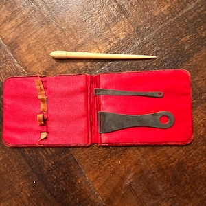 May include: A red folding case with a fabric interior containing two flat metal tools with circular holes at one end. A separate, smooth, light-colored pointed tool rests above the open case. The case features three small fabric loops on the left side.