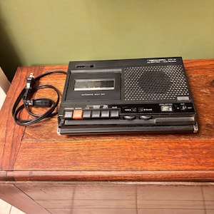May include: A black Realistic CTR-81 cassette recorder featuring a top-loading tape compartment, a built-in speaker, and a series of control buttons including record, play, rewind, fast forward, and pause. It includes an attached black power cord.