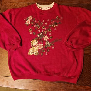 May include: A long-sleeve red sweatshirt featuring a crew neckline and a graphic print on the front. The design depicts a light-colored kitten wearing a red and white checkered bow, surrounded by branches with pink flowers, green leaves, and birds.