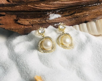 May include: A pair of gold earrings with a pearl and clear crystal accents. The earrings are on a white sandy surface.