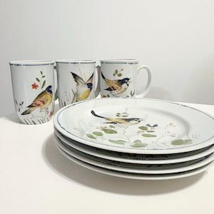 May include: A set of white ceramic tableware featuring hand-painted bird designs. The set includes three mugs and four plates. The plates and mugs have a blue trim. The birds are depicted in various poses with colorful plumage, surrounded by floral and botanical elements.