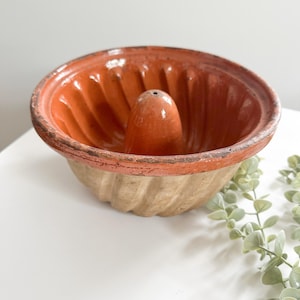 May include: An antique, terracotta-colored bundt pan with a central cone. The pan has a ribbed interior and a light beige exterior. The rim is a darker shade of terracotta, showing signs of age. A classic kitchenware item.