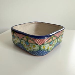 May include: Square ceramic bowl with a hand-painted design. The rim is dark blue, and the exterior features a repeating pattern of red, green, and blue floral motifs. The bowl has a light beige interior and is suitable for serving or display.