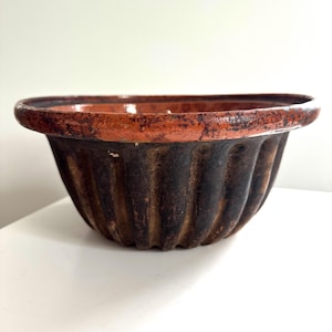May include: A vintage, fluted bowl with a wide, rounded rim. The bowl features a dark brown exterior with vertical grooves and a reddish-brown rim. The bowl is likely made of ceramic or earthenware and sits on a white surface.