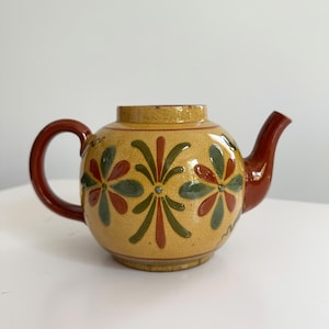 May include: A round ceramic teapot with a yellow crackle glaze finish. The body features a repeating pattern of stylized flowers in dark red and olive green. It has a solid dark red handle and a matching curved spout, with no lid present.