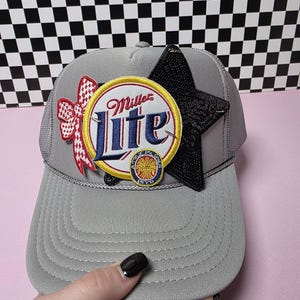 Può includere: Cappellino da camionista grigio con il logo Miller Lite su una toppa circolare con dettagli rossi e gialli, una stella nera e un fiocco rosso. Il cappello ha un retro in rete e una visiera curva.