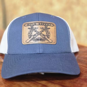 May include: A blue and white baseball cap featuring a rectangular tan leather patch on the front. The patch is engraved with the text RIFLE EXPERT above an illustration of two crossed rifles surrounded by a wreath and anchor emblem.