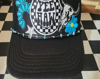 Może przedstawiać: Czarna czapka typu trucker z dekoracyjnym wzorem. Czapka zawiera napis "Yee Haw" wewnątrz podkowy, czaszkę byka i turkusowe akcenty. Gwiazda i niebieski kwiat dopełniają projekt.