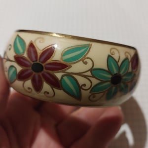 Könnte beinhalten: Cremefarbenes Armreif-Armband mit floralem Design. Das Armband hat weinrote und türkisfarbene Blumen mit schwarzen Zentren und goldenen Akzenten. Die Blumen sind vor einem cremefarbenen Hintergrund angeordnet. Das Armband hat eine breite, abgerundete Form.