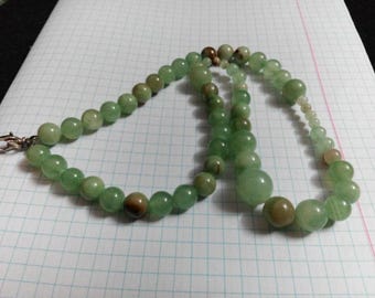 Puede incluir: Un collar compuesto por cuentas verdes y marrones de varios tamaños. Las cuentas están ensartadas, formando una forma circular. El collar descansa sobre una superficie blanca con un patrón de cuadrícula.