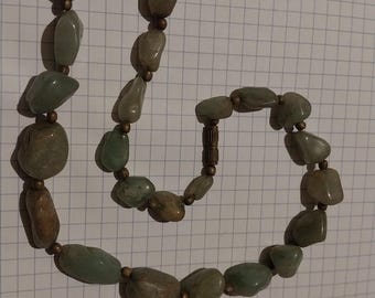 Puede incluir: Un collar con piedras de color verde claro y marrón de forma irregular, ensartadas con pequeñas cuentas doradas. El collar tiene un cierre y se muestra sobre un fondo de cuadrícula blanca.