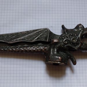 May include: A detailed, antique-style nutcracker in the shape of a dragon. The dragon is dark metallic with intricate detailing on the wings, head, and body. The nutcracker is designed to crack nuts.