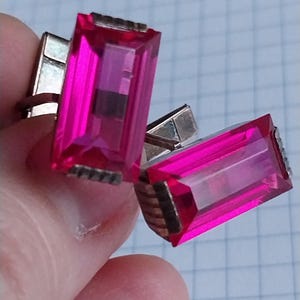 Puede incluir: Un par de gemelos plateados con grandes piedras preciosas rectangulares de color rosa intenso. Las piedras tienen un aspecto facetado y el engaste metálico presenta un diseño texturizado. Los gemelos se muestran sobre un fondo cuadriculado.
