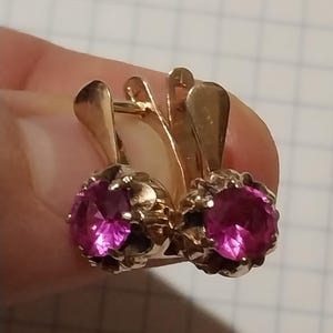 May include: A pair of gold earrings with pink gemstones. The earrings have a simple design with a round gemstone set in a gold setting.