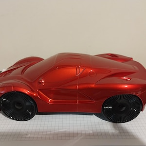 May include: A shiny red toy car with black wheels. The car has a sleek, aerodynamic design, with a low profile and curved lines. The wheels have a unique, spoked design. The car is sitting on a white grid.