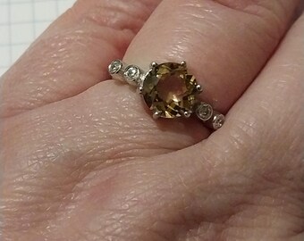 May include: A silver ring featuring a large, round, light brown gemstone as the centerpiece. The ring is accented with smaller, clear gemstones. The ring is a piece of jewelry.