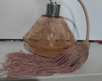 Può includere: Un flacone di profumo vintage con un corpo in vetro color pesca, inciso con disegni floreali. È dotato di un tappo in metallo argentato e un nappa decorativa. L'altezza del flacone è di circa 13 cm.