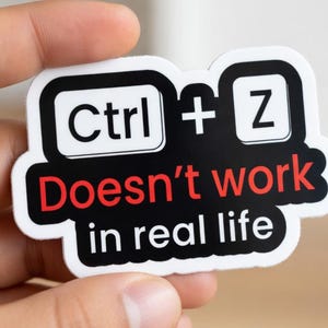 Könnte beinhalten: Ein weißer Aufkleber mit schwarzem Rand mit dem Text "Ctrl + Z Doesn't work in real life" in einem Tastaturtasten-Design. Der Aufkleber wird in einer Hand gehalten.