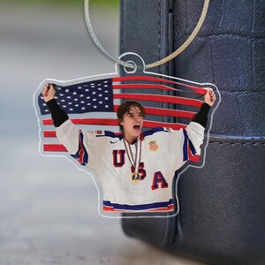 Puede incluir: Llavero acr&iacute;lico transparente con un jugador de hockey sobre hielo con una camiseta blanca con detalles rojos y azules, las letras "USA" y una medalla de oro. El fondo es la bandera americana.