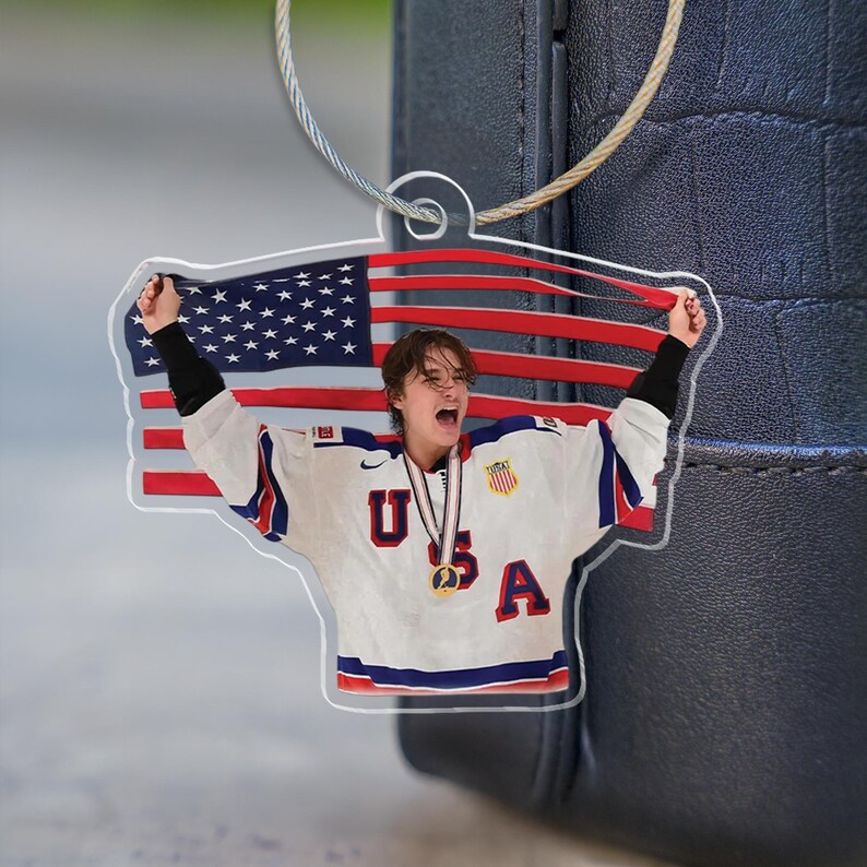 Puede incluir: Llavero acr&iacute;lico transparente con un jugador de hockey sobre hielo con una camiseta blanca con detalles rojos y azules, las letras "USA" y una medalla de oro. El fondo es la bandera americana.