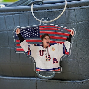 Puede incluir: Llavero acr&iacute;lico transparente con una persona con una camiseta de hockey de EE. UU. sosteniendo una bandera estadounidense. La camiseta tiene las letras "USA" y una medalla de oro. El llavero est&aacute; sujeto a una cadena plateada.