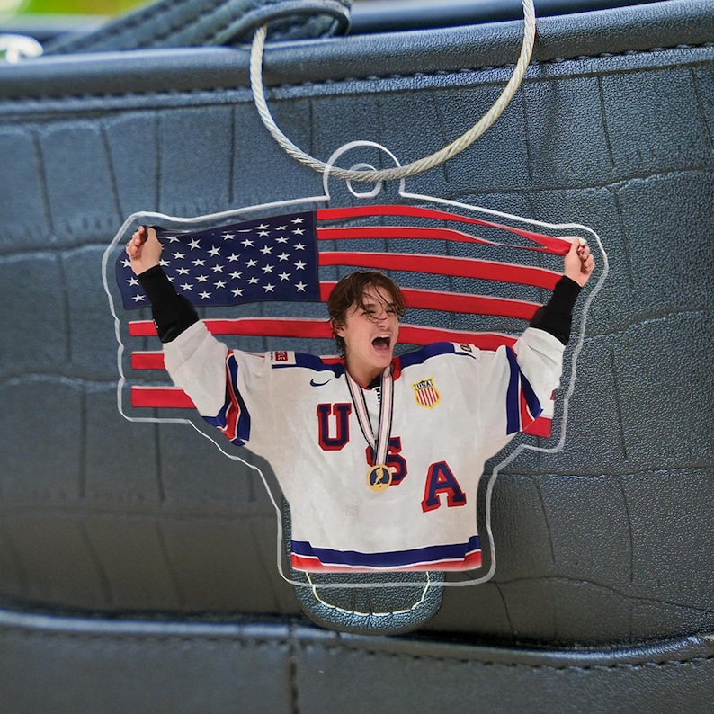 Puede incluir: Llavero acr&iacute;lico transparente con una persona con una camiseta de hockey de EE. UU. sosteniendo una bandera estadounidense. La camiseta tiene las letras "USA" y una medalla de oro. El llavero est&aacute; sujeto a una cadena plateada.