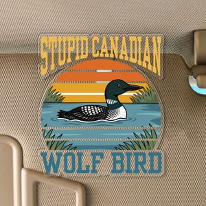 May include: A clear plastic car visor featuring a cartoon of a loon swimming in a lake with a sunset background. The text reads "STUPID CANADIAN" above and "WOLF BIRD" below the image.