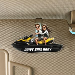 Puede incluir: Un clip de visera de moto acuática negro y amarillo con una pareja de dibujos animados. La moto acuática tiene el texto "DRIVE SAFE BABY" en blanco. El clip está sujeto a una visera de coche beige.