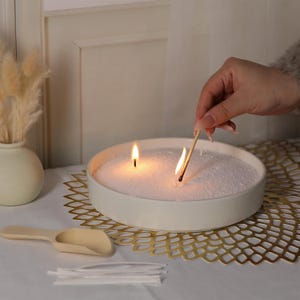 Puede incluir: Un cuenco de cer&aacute;mica blanca lleno de arena blanca y dos velas encendidas. Una mano sostiene una cerilla para encender la segunda vela. El cuenco est&aacute; sobre un encaje de metal dorado.
