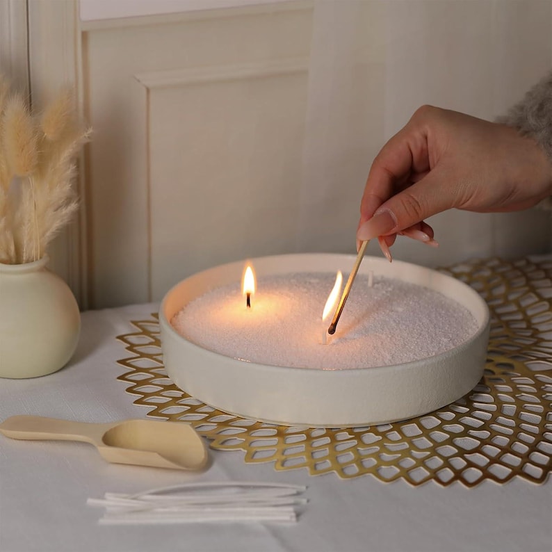 Puede incluir: Un cuenco de cer&aacute;mica blanca lleno de arena blanca y dos velas encendidas. Una mano sostiene una cerilla para encender la segunda vela. El cuenco est&aacute; sobre un encaje de metal dorado.
