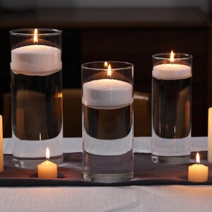 Puede incluir: Tres jarrones cil&iacute;ndricos de vidrio transparente con velas flotantes. Las velas son blancas y tienen una peque&ntilde;a llama. Los jarrones est&aacute;n llenos de agua y las velas flotan en la superficie del agua.