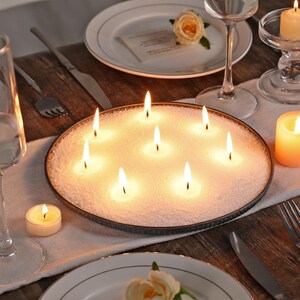 Puede incluir: Una bandeja redonda de metal llena de arena blanca y nueve peque&ntilde;as velas encendidas. La bandeja est&aacute; sobre una mesa de madera con un mantel blanco y un plato con una flor.