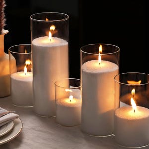 Puede incluir: Cinco portavelas de vidrio con velas blancas encendidas en su interior. Las velas son de diferentes tama&ntilde;os y est&aacute;n dispuestas en fila sobre una superficie blanca.