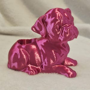 May include: A pink 3D printed pug figurine. The figurine is lying down with its head turned to the side. It has a smooth, glossy finish.