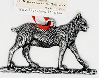 May include: A silver pewter ornament of a bobcat walking on a branch. The ornament is handmade in Montana and is part of the American Wildlife Series. The ornament has a red ribbon attached to it.