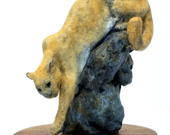 May include: A cougar sculpture in a descending pose, atop a dark rock formation, mounted on a wooden base. The sculpture is tan and brown, with a bronze plaque that reads "SILENT DESCENT" by Eric Thorsen.