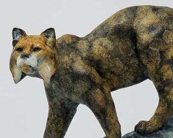 May include: A miniature bobcat sculpture by Eric Thorsen. The bobcat is depicted in a walking pose, perched on a weathered log. The sculpture has a brown and tan spotted coat, with a wooden base and a brass nameplate.