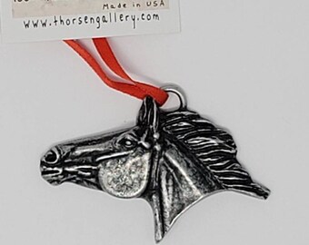 May include: Silver horse head ornament with a red ribbon. The ornament is part of the Frontier America American Wildlife Series, 100% handmade in Montana, USA. The tag reads "Frontier America American Wildlife Series 100% Handmade in Montana Made in USA www.thorsengallery.com"