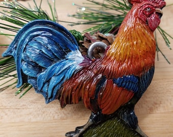 May include: A colorful rooster ornament with a blue, white, and red tail, a red comb, and a yellow and orange body. The ornament has a silver loop for hanging.