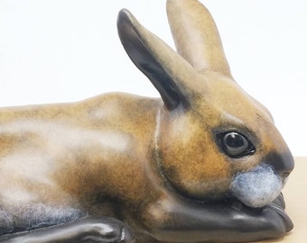 May include: A ceramic sculpture of a brown rabbit lying down with its head turned to the side. The rabbit has black eyes and a white patch on its nose.