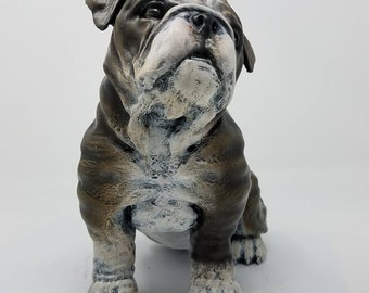 May include: A brown and white bulldog figurine with a wrinkled face and a short tail. The figurine is sitting on a white surface.
