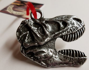 May include: Silver-toned Tyrannosaurus Rex skull ornament with a red ribbon attached. The ornament is detailed with a textured surface and a loop for hanging.