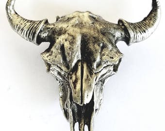 May include: A silver-colored, detailed sculpture of a bull skull with prominent horns. The skull has a weathered appearance, with visible ridges and textures. The horns curve upwards and outwards, adding to the skull's imposing presence. The sculpture is likely a decorative item.
