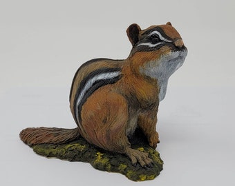 May include: A realistic figurine of a chipmunk with brown and black fur, sitting on a green base. The chipmunk has a white stripe down its back and a white patch on its chest.
