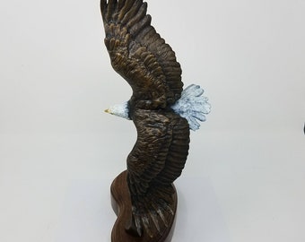 May include: A bronze sculpture of a bald eagle in flight, with its wings spread wide and its head turned to the side. The eagle is perched on a wooden base.
