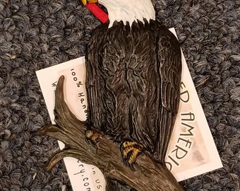 May include: A hand-painted bald eagle ornament perched on a branch. The eagle is painted in shades of brown, black, and white with yellow accents. The ornament is made in the USA and is 100% handmade.