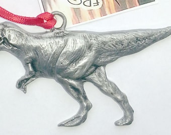 May include: A silver-colored pewter Tyrannosaurus Rex dinosaur ornament. The ornament is hanging from a red ribbon.