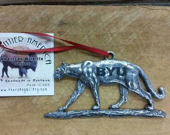 May include: A silver pewter ornament of a cougar walking on a log. The ornament has the text "BYU" engraved on the cougar's side. The ornament is part of the Frontier America American Wildlife Series.
