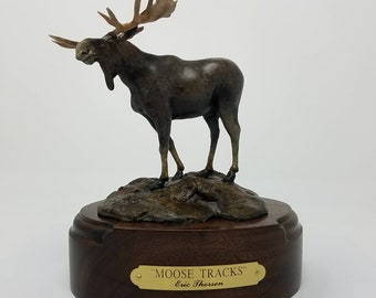 May include: Bronze sculpture of a moose standing on a rocky base. The sculpture is on a wooden base with a brass plaque that reads "MOOSE TRACKS" Eric Thorsen.